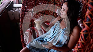 Relaxed young woman using a smartphone and drinking coffee with ice while sitting in hanging wicker chair at cafe