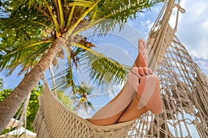 Relaxing beach vacation with woman`s feet in hammock between coconut palm tree. Exotic tropical island hotel resort. Sunny warm