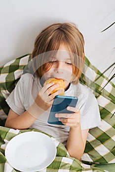 Relaxing in bed, a 9-10-year-old boy with flowing hair indulges in a toast, engrossed in his mobile phone, exploring