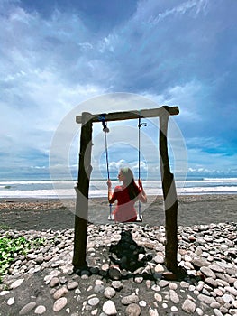 Relaxing day at Medewi beach, young girl in red dress on the swing, Bali, Indonesia. Black sand beach.