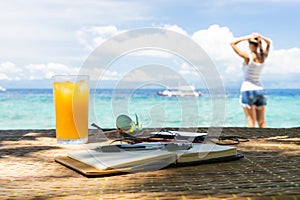Relaxing girl, opened empty notepad is on the table with sunglasses, phone and headphones at the tropical sea background