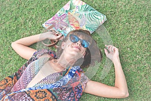 Relaxing in grass. Top view of beautiful young woman in sunglasses and pareo lying on the green grass with beach bag in