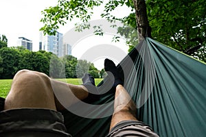 relaxing in the green hammock in the summer in a city park. Socks in feet close up. Buildings and grass background. Man legs