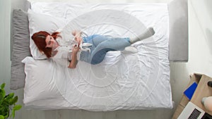 Relaxing at home, young woman uses a smartphone to browse internet, orders goods online while lying on bed, top view