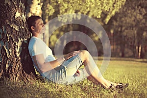 Relaxing man sitting under a tree with eyes closed meditating enjoying the warm evening sunset in profile