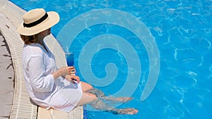 relaxing senior woman dangling her legs in an outdoor swimming pool holding a blue refreshing cocktail wearing a straw