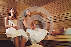 Relaxing teens couple wrapped white sheets lying and sweating on wooden bench in Hot Finnish sauna enjoying pleasant body care
