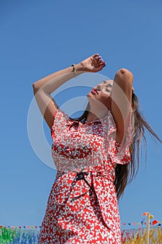 Relaxing young millennial woman in red dress against blue sky. Beautiful smiling girl on summer having fun. Enjoying sun