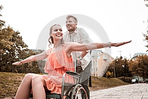 Reliable man smiling and pushing the wheelchair of his happy girlfriend