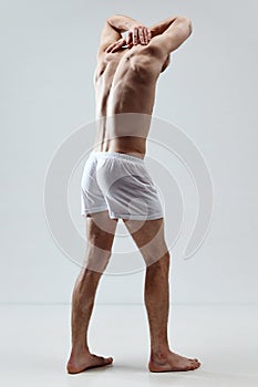 Relief back. Full-length portrait of handsome young man with blonde hair, muscular body in underwear against grey studio