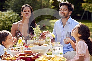 Religious Family Saying Prayers Or Grace Before Eating Outdoor Meal In Garden