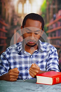 Religious man sitting and holding rosary looking down, red book lying on desk in front, religion concept