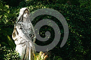 Religious sculpture of nun stone material figure in parkland environment with with green blurry foliage of bushes