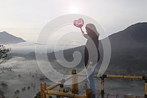 Religious woman holding red love sign in hand with text God against the top of mountain in the misty morning.