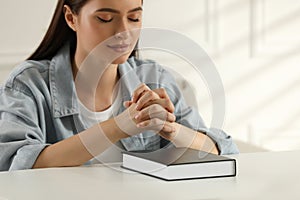 Religious young woman praying over Bible at table indoors