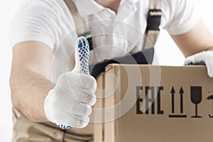 Relocation services concept. Loader in uniform shows thumb up closeup. Mover with cardboard box. Delivery man carrying moving box