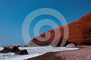 Remaining red sandstone stone arch in Legzira Beach. Rugged coastline in Tiznit Province of Morocco, Africa. Atlantic Ocean waves.