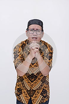 A remorseful man begging and saying sorry. Asking for a second chance. Wearing a batik shirt and songkok skull cap with hand on