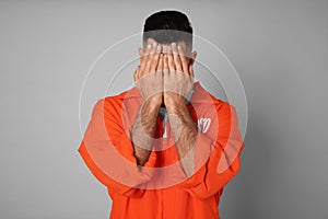 Remorseful prisoner in orange jumpsuit hiding his face on grey background