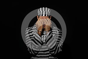 Remorseful prisoner in striped uniform with handcuffs hiding his face on black background