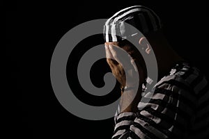 Remorseful prisoner in striped uniform with handcuffs hiding his face on black background, space for text