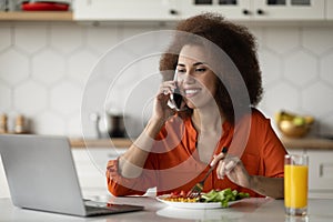 Remote Business. Black Woman Using Cellphone And Working On Laptop In Kitchen