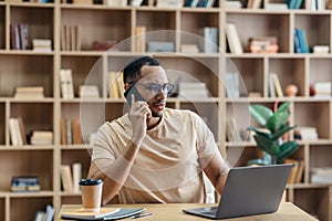 Remote business concept. Concerned arab man talking on cellphone and using laptop while sitting at desk at home