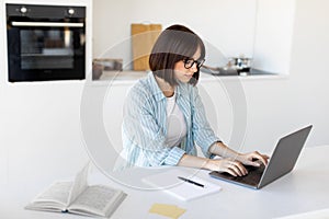 Remote business. Young freelancer lady working on laptop in kitchen, sitting at table and typing on pc keyboard