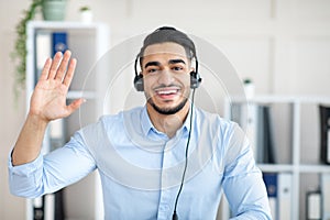 Remote communication concept. Happy Arab guy with headset having video call at office, waving at webcam