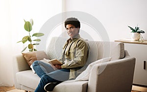 Remote education concept. Joyful African American student reading book on comfy couch at home