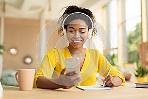 Remote learning. Cheerful black lady in wireless headphones sitting at desk using smartphone and writing in her notebook
