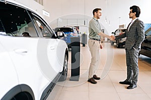 Remote side view of confident car salesman in business suit and cheerful male customer in auto dealership. Two happy