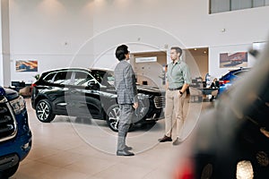 Remote view of doubtful male customer buying car in showroom talking to sales agent in business suit looking at auto
