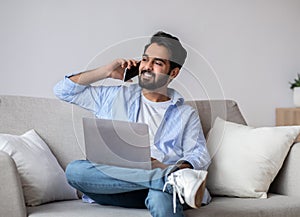 Remote Work. Arab Man Using Laptop And Talking On Cellphone At Home