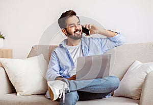 Remote Work. Arab Man Using Laptop And Talking On Cellphone At Home