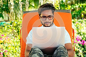 Remote work and freelancing.young indian man sitting orange lounger deck chair with laptop on beach.freelancer surfing