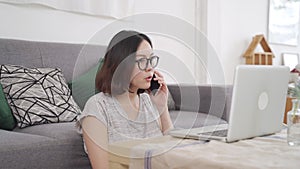 Remote working at home office at afternoon, happy Asian woman sitting on floor working desk in living room, typing computer laptop