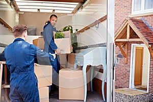 Removal Company Workers Unloading Furniture And Boxes From Truck Into New Home On Moving Day