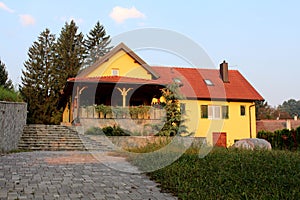 Renovated yellow suburban family house with large porch in front and stone tiles driveway surrounded with high uncut grass