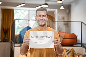 Reopening office safely. Young happy man showing paper with text SOCIAL DISTANCE at camera and smiling, standing in the