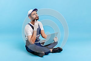 Repair service, online help. Portrait of pensive handyman in workwear sitting cross-legged with laptop