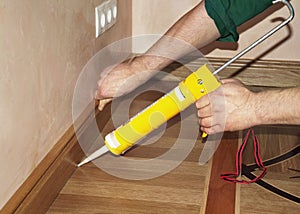 Repairman Installing Skirting Board Oak Wooden Floor with Caulking Gun Glue from Cartridge.