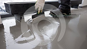 Repairman in protective gloves and uniform using metal spatula for aligning liquid self-levelling floor screed. Worker