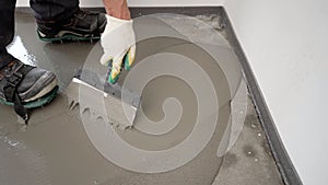 Repairman in protective gloves using a metal spatula to level a liquid self-levelling floor screed. A worker pours