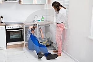 Repairman Repairing Pipe While Woman In The Kitchen