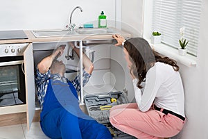 Repairman Repairing Pipe While Woman In The Kitchen