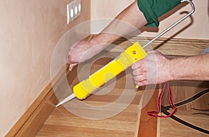 Repairman's hands Installing Skirting Board Oak Wooden Floor with Caulking Gu