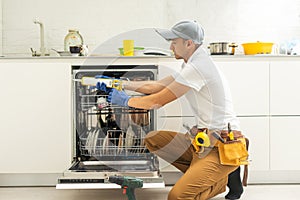 Repairman in uniform repairs dishwasher in kitchen. Young man specialist unscrews parts with screwdriver checking state