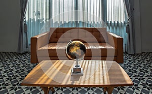 Replica globe on wooden coffee table front to brown leather sofa on background of white curtains and glass windows in living room