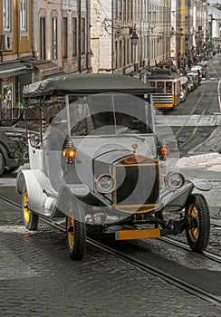 Replica of an old gray electric tourist car going up the slope of a cobblestone street with tram tracks through the old Alfama
